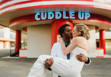bride, White, and groom, Black, kiss in front of Glen Echo Park Cuddle Up Pavilion