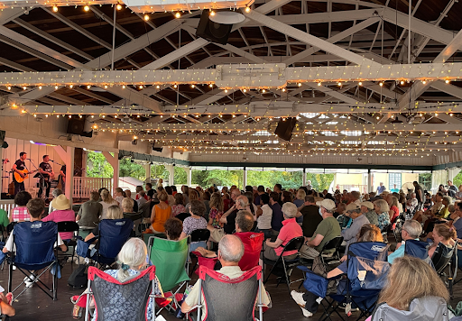 A full house attending a concert in the Park's Bumper Car Pavilion with people sitting on benches and twinkling lights on the ceiling.